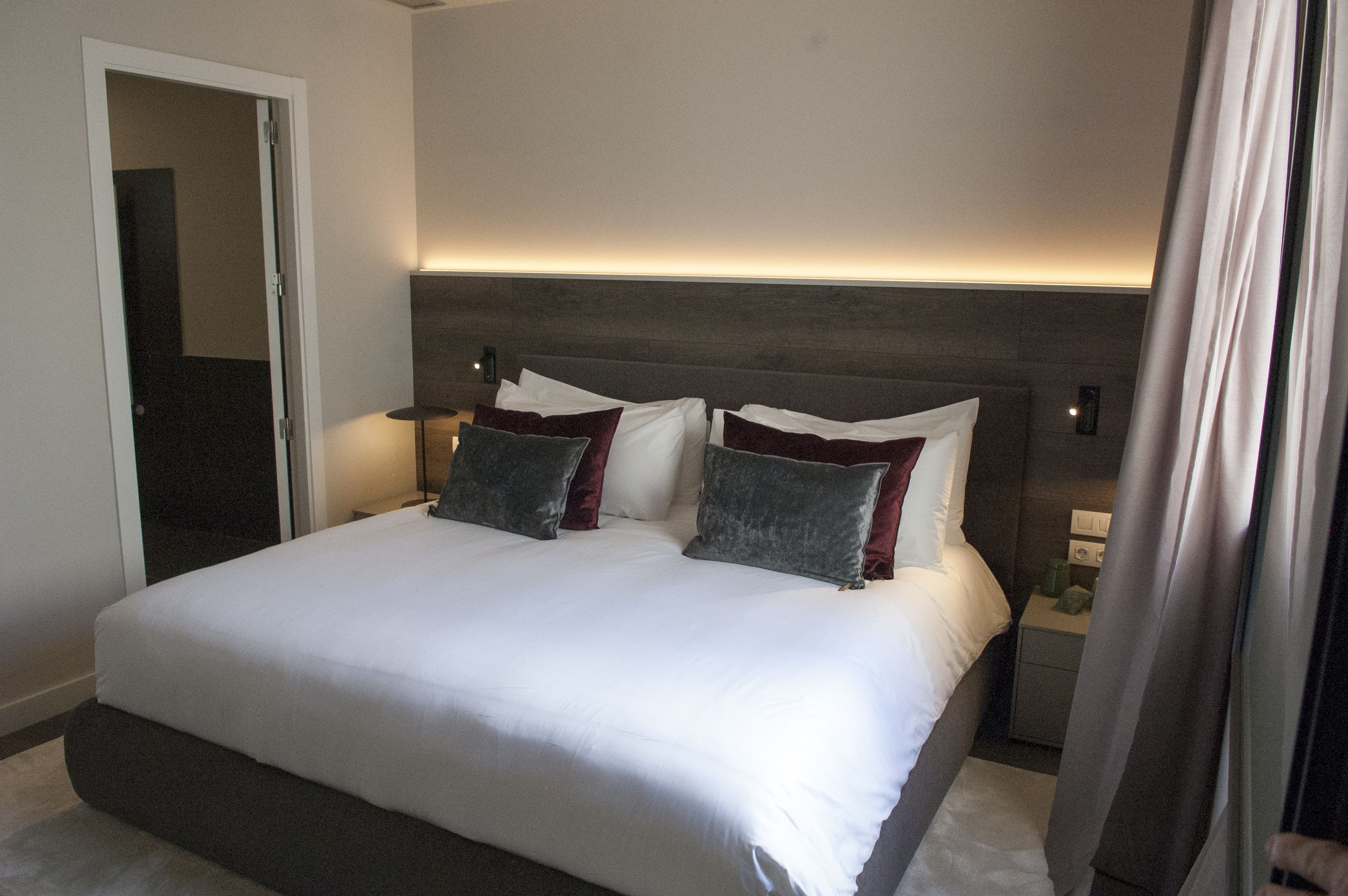 En pænt redt seng med hvide sengetøj og to puder, et natbord med lampe, og en moderne sengegavl.