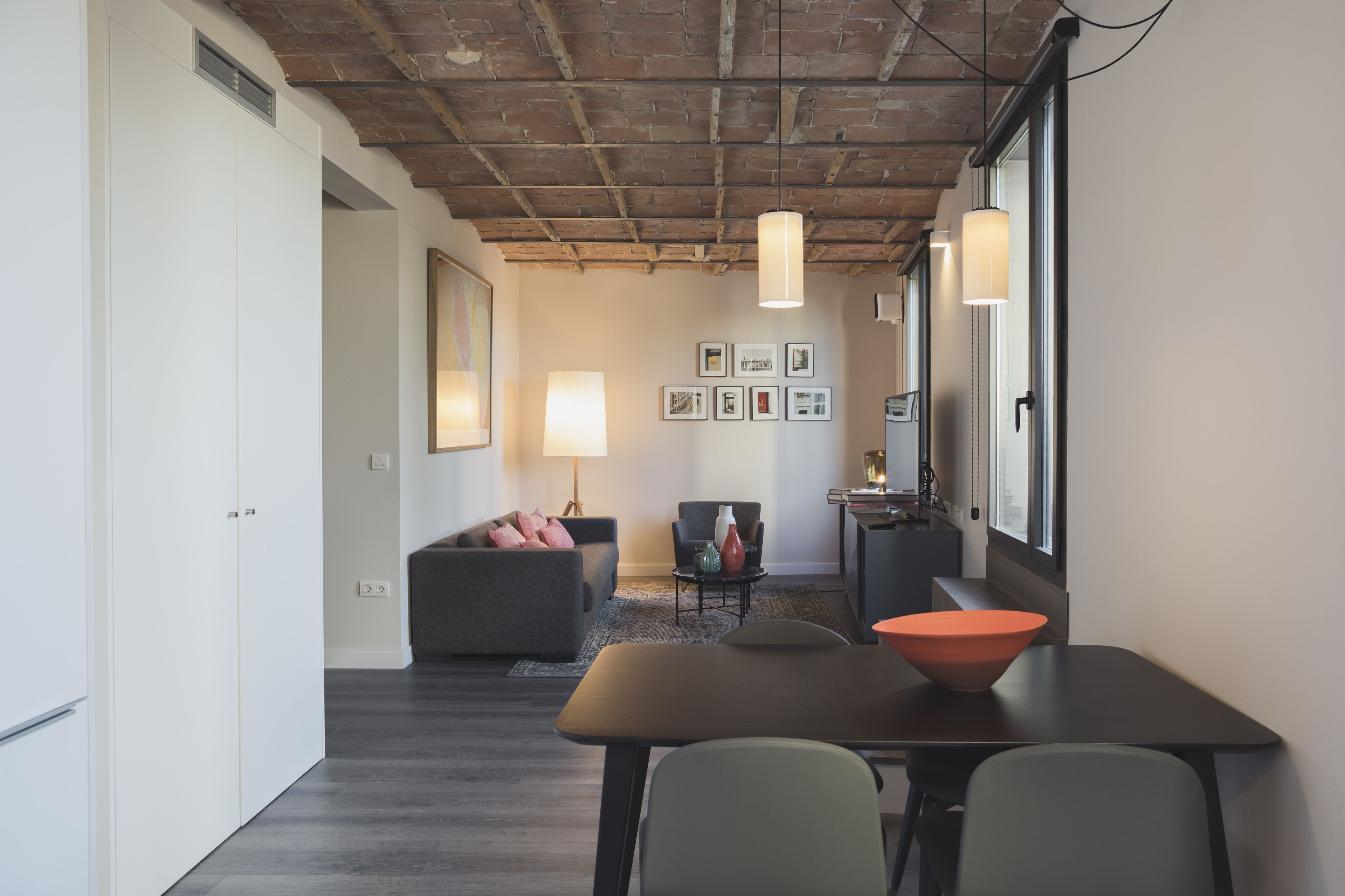 A modern living room with a dark wooden table, a grey sofa, and a black armchair. There is a large window on the right side and a door on the left.