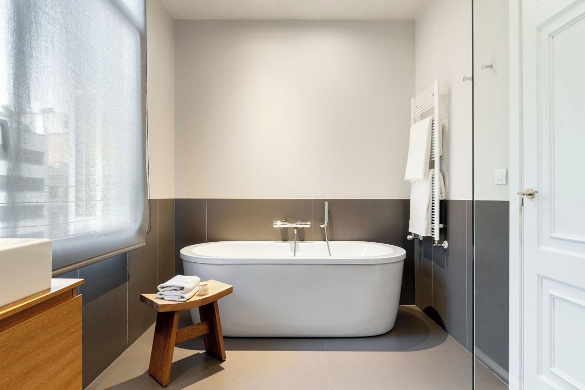 Een moderne badkamer met een vrijstaand bad, een houten kruk met handdoeken en een groot raam.
