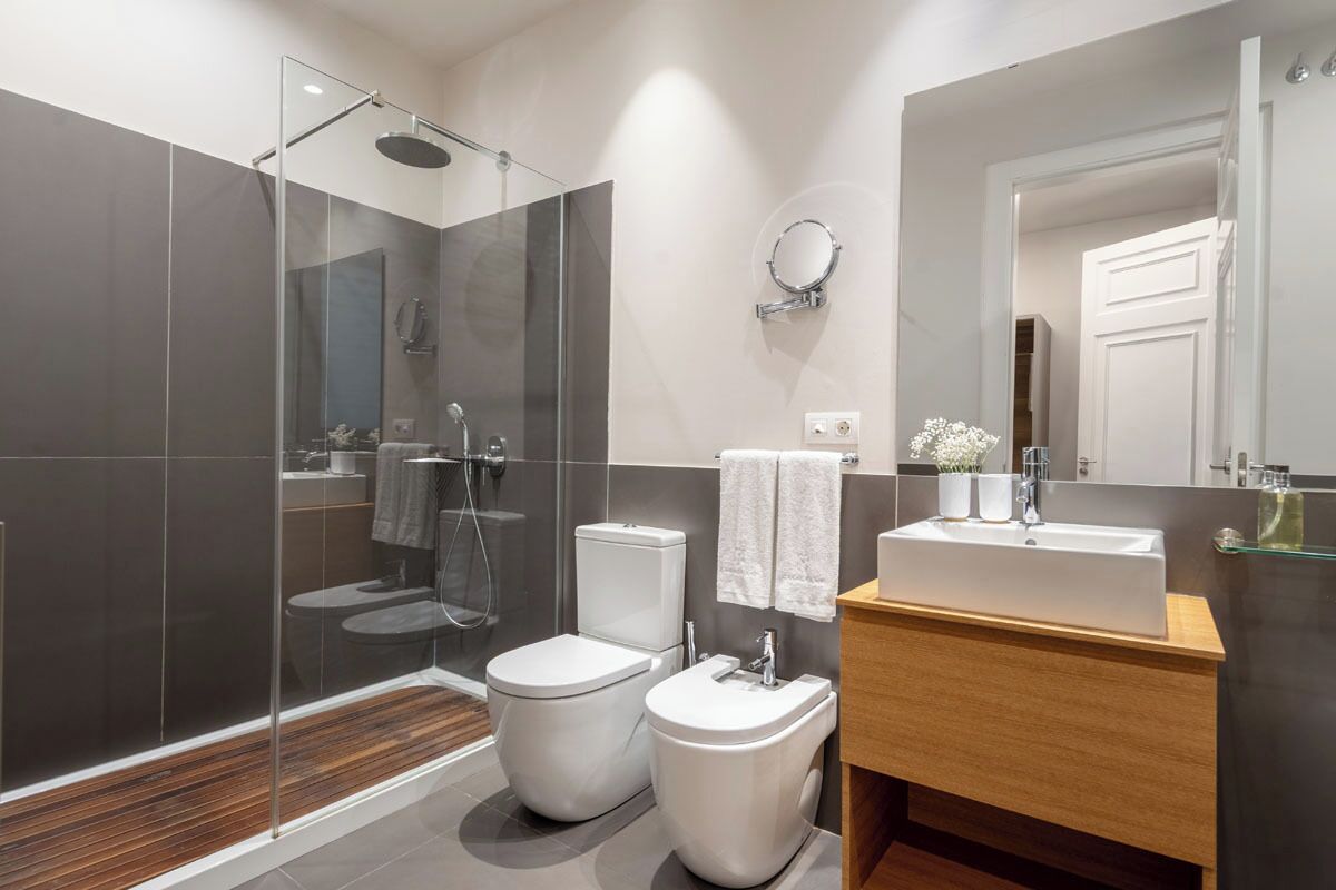 A modern bathroom with a glass-enclosed shower, two toilets, a sink, and a mirror.