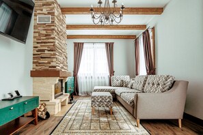 Ein Wohnzimmer mit Kamin aus Stein, Holzbalken, einer grünen Anrichte, einem gemusterten Sofa und einem großen Fernseher.
