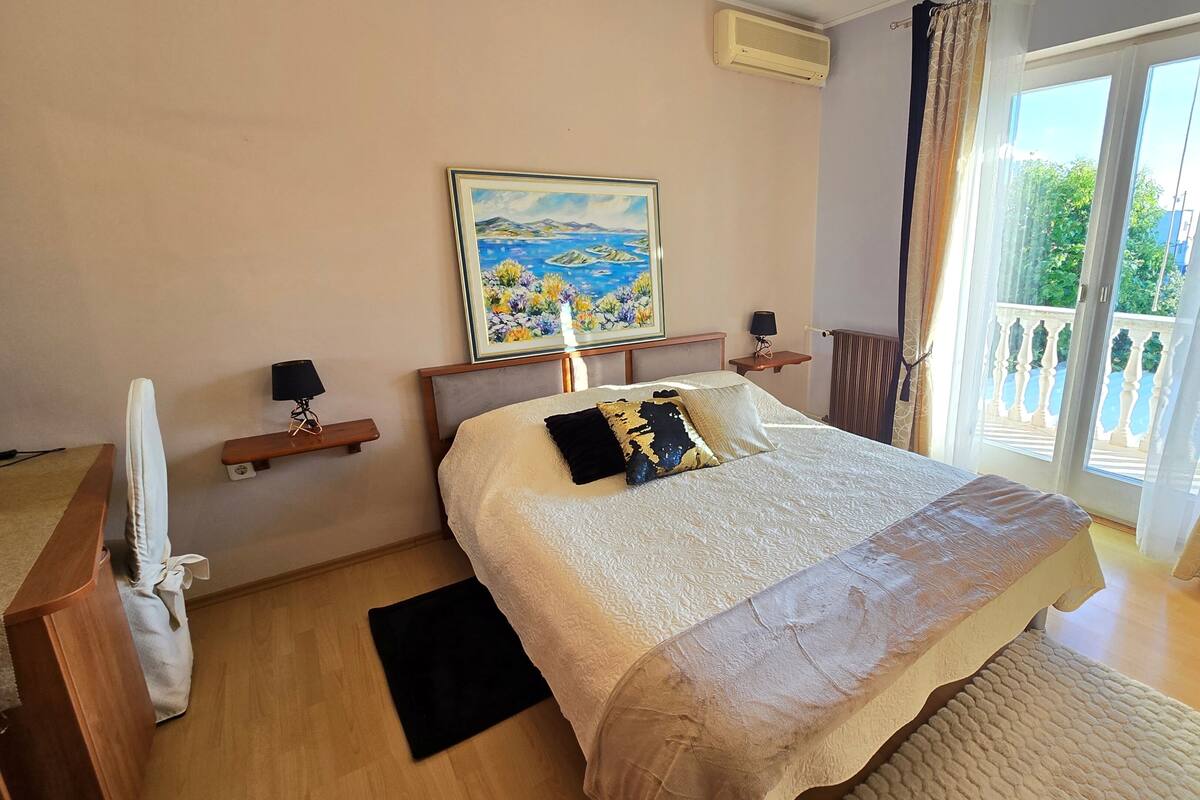 A bedroom with a bed, a painting, a window with curtains, a bedside table, and a radiator.