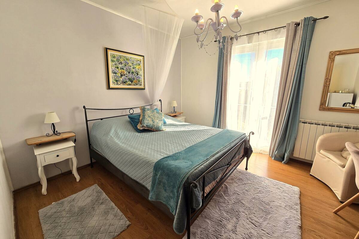A bedroom with a bed, bedside tables, a nightstand, a chair, a window with curtains, a chandelier, and a radiator.