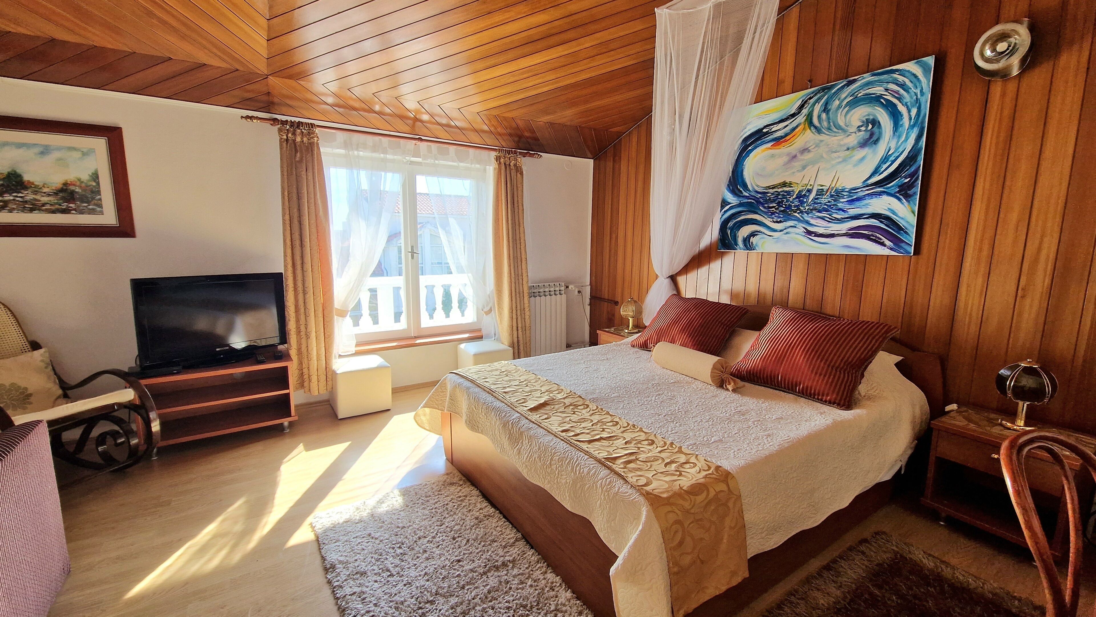 A bedroom with a wooden ceiling, a bed with a patterned bedspread, a painting above the bed, a television on a stand, and a window with curtains.