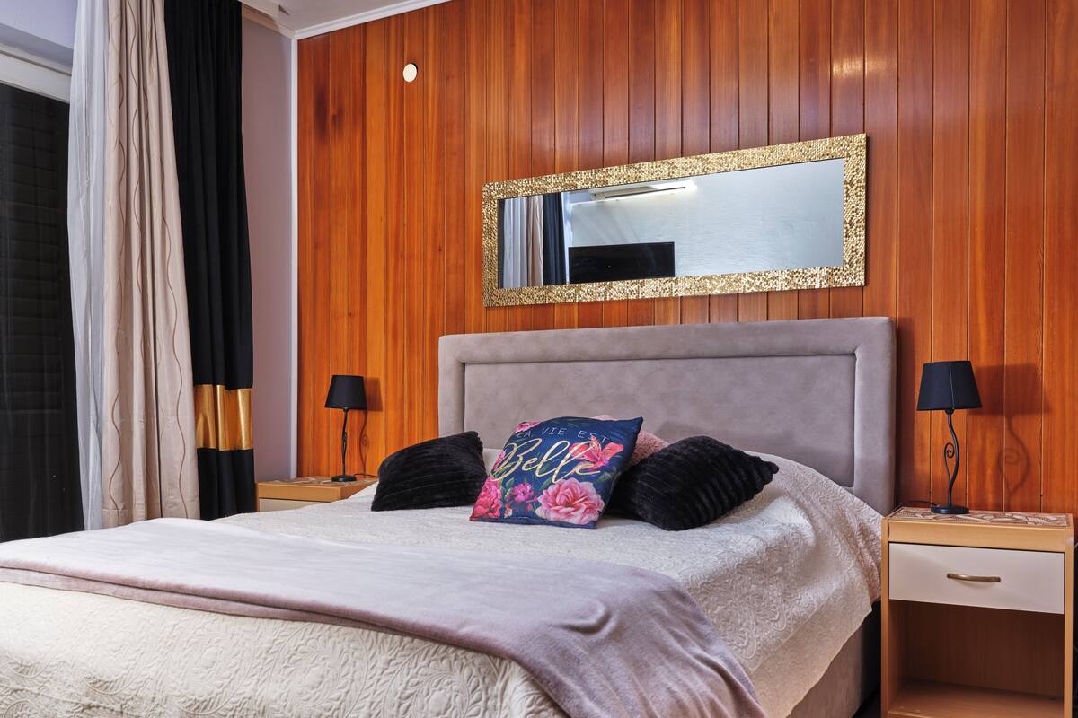 A bedroom with a bed, two bedside tables, a nightstand, a mirror, and a window with curtains.