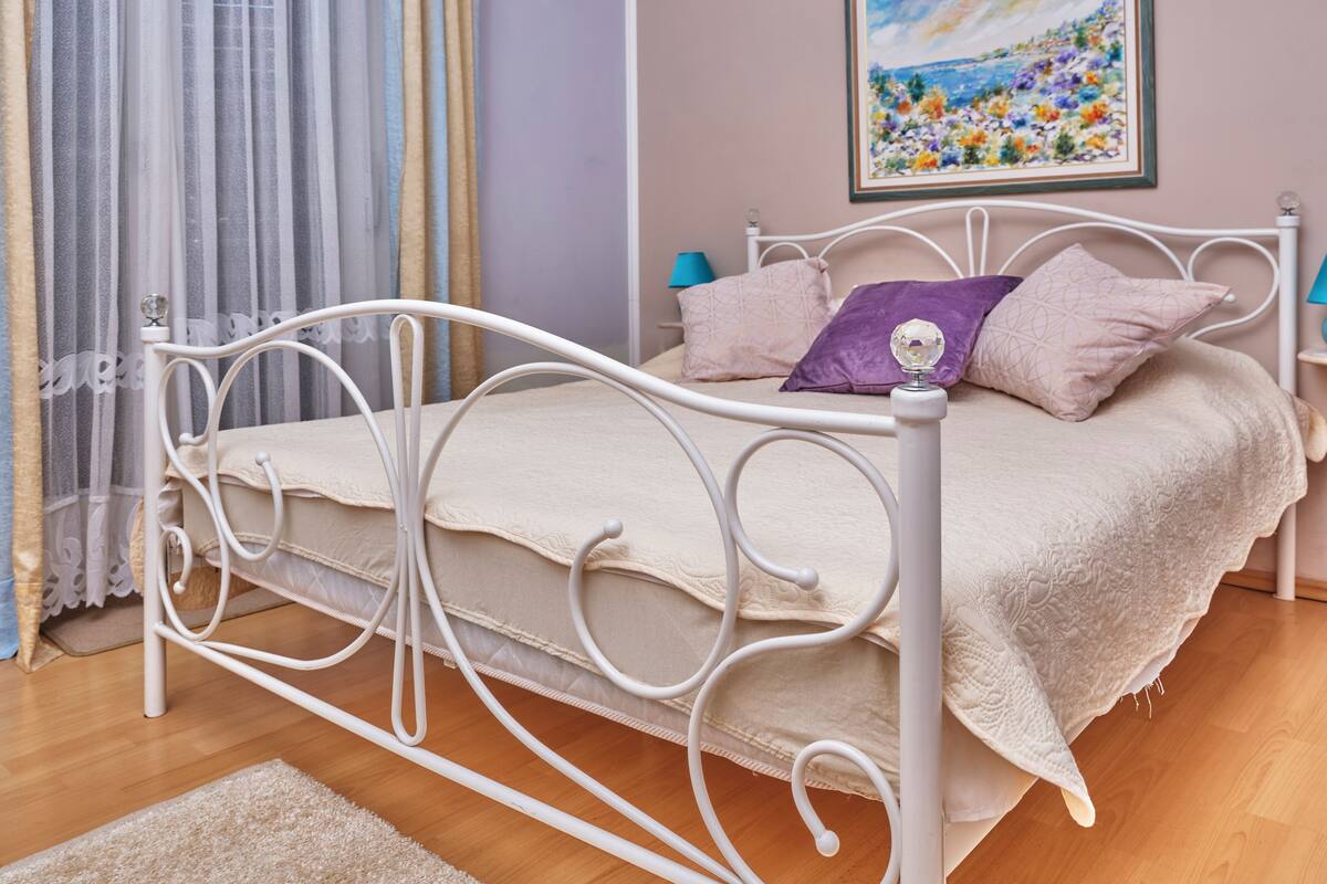 A white metal bed with a beige bedspread and pillows, a wooden floor, a beige rug, a painting on the wall, and a window with curtains.