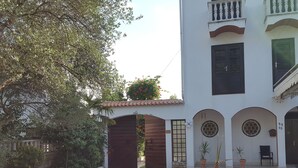 Um pátio com piso de azulejos, portão de madeira, plantas em vasos e um edifício com telhado e portas em arco.