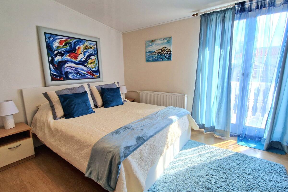 A bedroom with a bed, a nightstand, a lamp, a painting, a radiator, and a window with curtains.