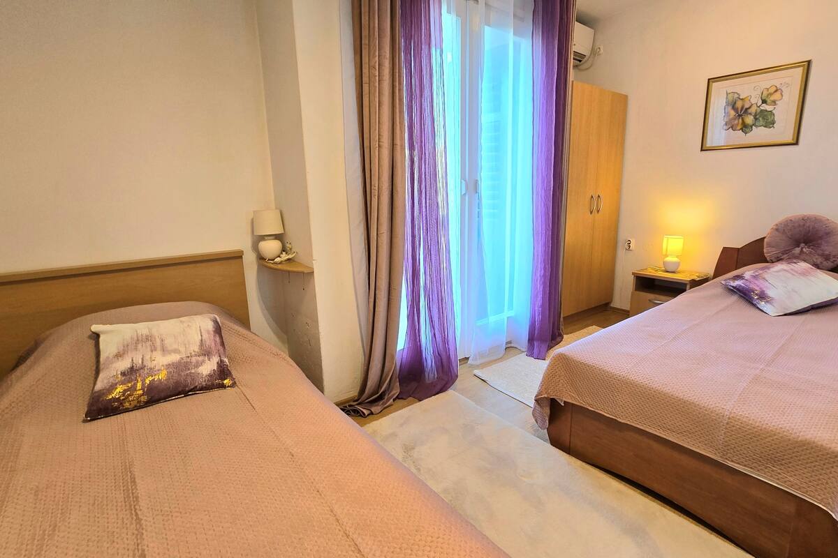 A hotel room with two beds, a wooden wardrobe, a small table with a lamp, and a framed picture on the wall.