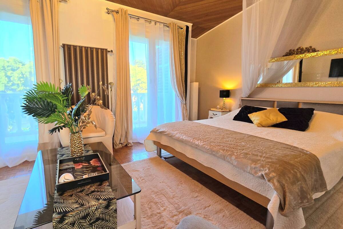 A bedroom with a bed, a nightstand, a lamp, a mirror, a window with curtains, and a plant on a table.
