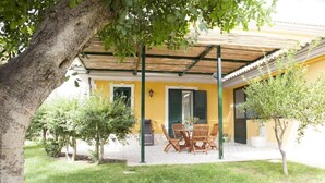 A covered patio with a table and chairs, a small garden with trees, and a yellow building with green shutters. - Valleforno Country House di Charme (Ispica)