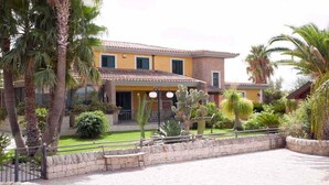 A two-story building with a yellow facade and a tiled roof. There are palm trees and cacti in the garden area. - Valleforno Country House di Charme (Ispica)