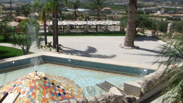 A swimming pool with a colorful mosaic dome, surrounded by palm trees and a view of a distant town. - Valleforno Country House di Charme (Ispica)