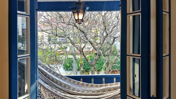 A hammock hanging from a balcony with a view of trees and a building in the background. - Farfalla Guest House (Rio de Janeiro)