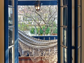 A hammock hanging from a balcony with a view of trees and a building in the background. - Farfalla Guest House (Rio de Janeiro)