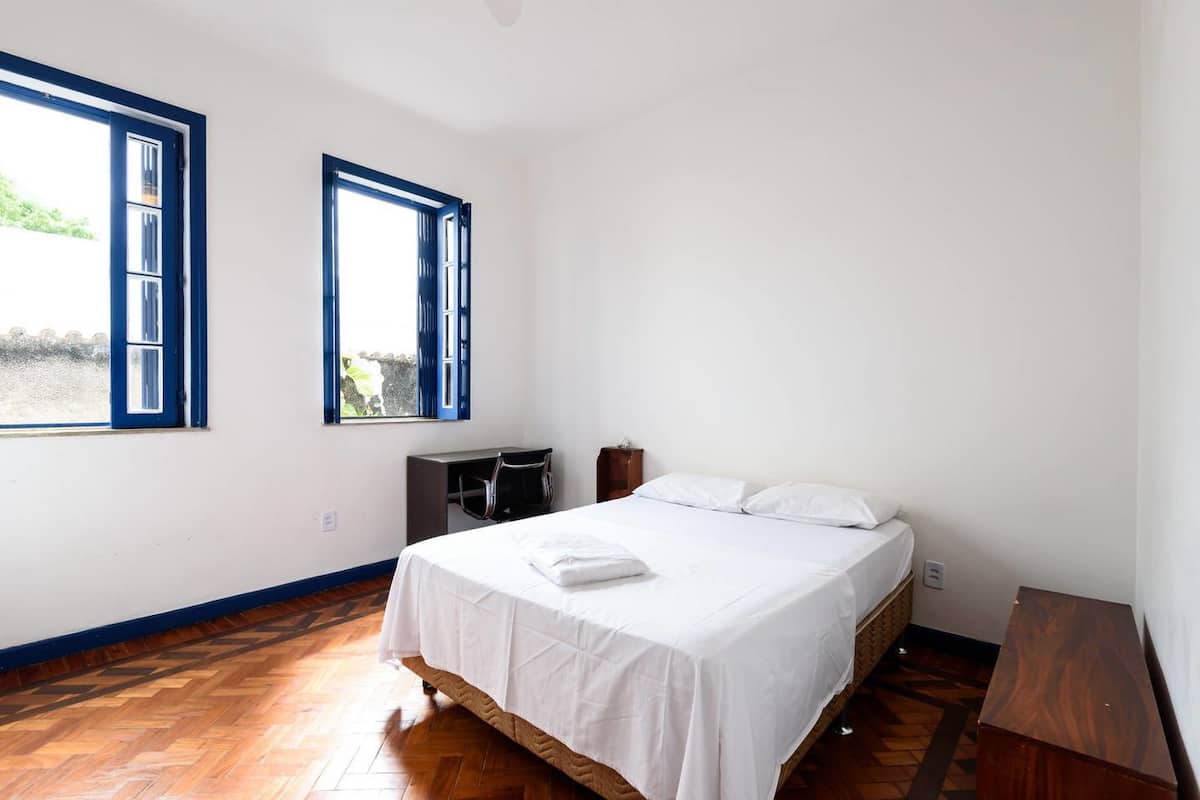 Un dormitorio con una cama, dos ventanas, un escritorio y una silla.