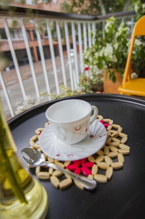 A white cup and saucer on a tray with a spoon and some pills, placed on a balcony with a railing and some greenery in the background.