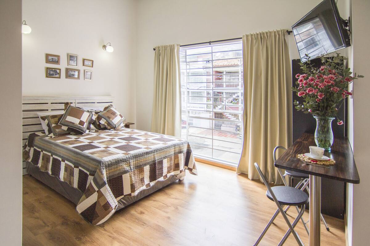 A bedroom with a bed, a desk with a chair, a window with curtains, and a wall-mounted TV.