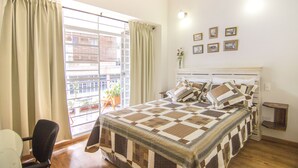 A bedroom with a large bed, a window with curtains, a small table, and a chair. - Jovita House BnB (Cali)