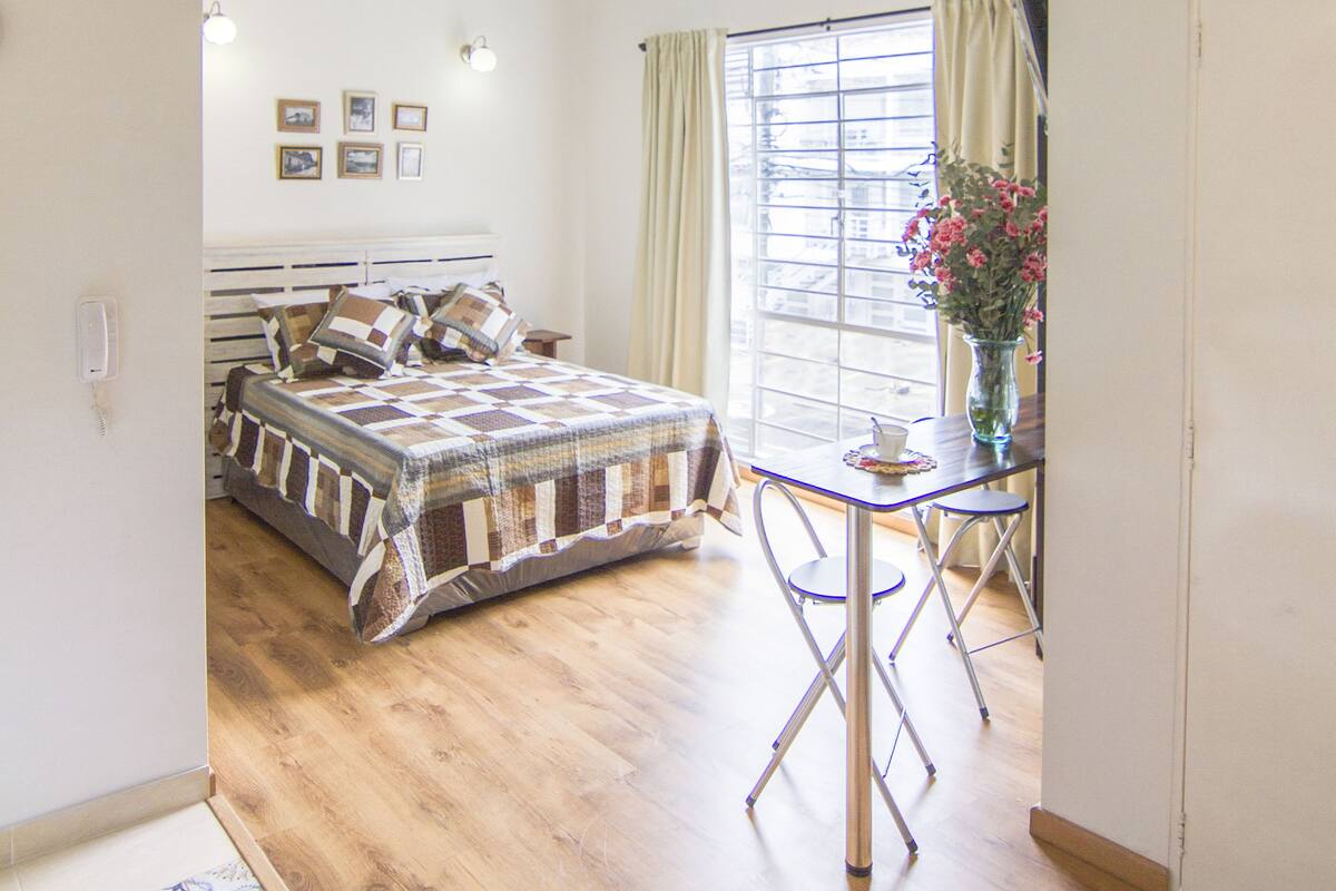 A bedroom with a bed, a small table with a vase, and a window with curtains.
