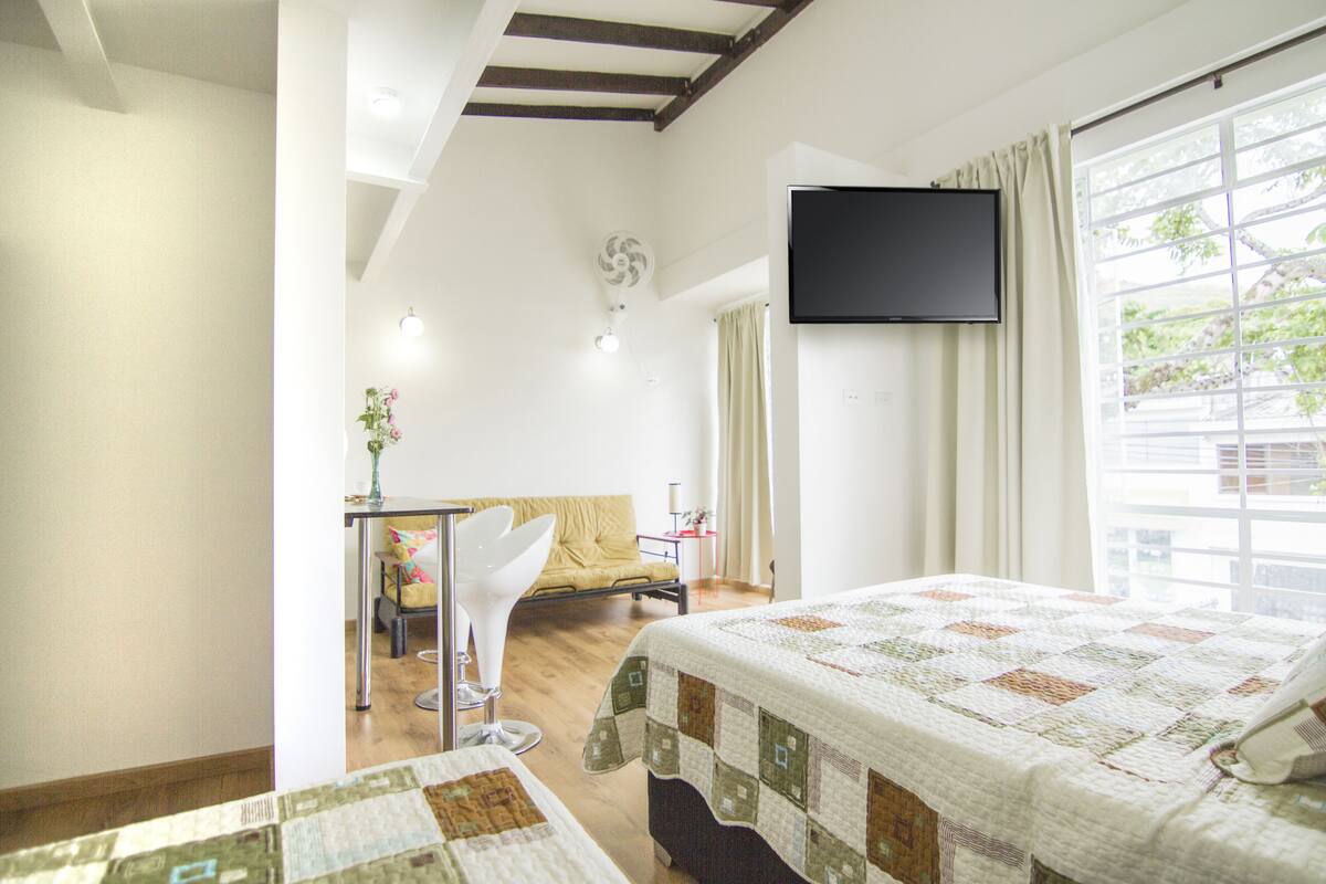 A hotel room with a bed, a TV mounted on the wall, a window with curtains, a chair, and a small table with a vase.