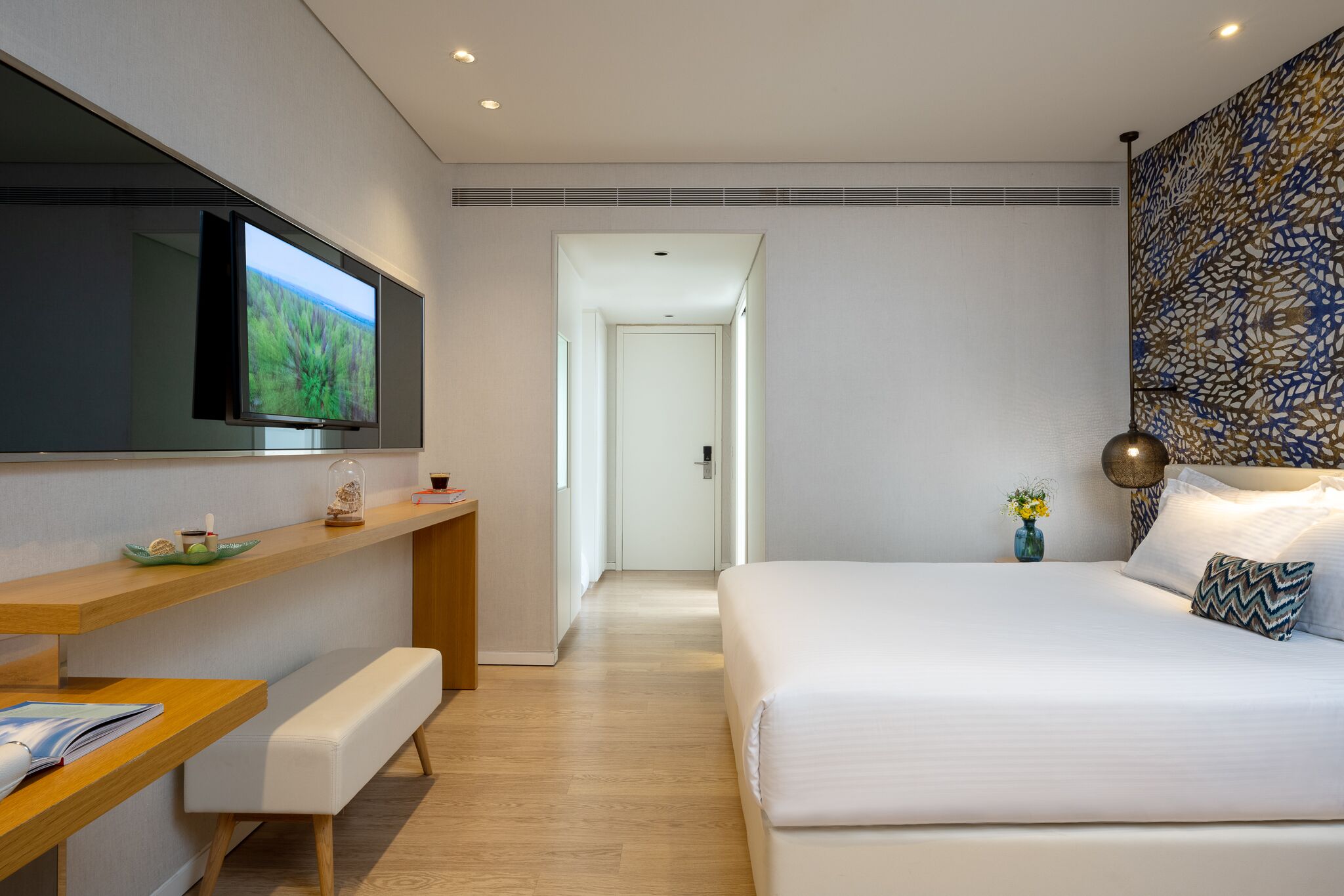 A modern hotel room with a large bed, a flat-screen TV mounted on the wall, a wooden desk with a bench, and a decorative wall panel.