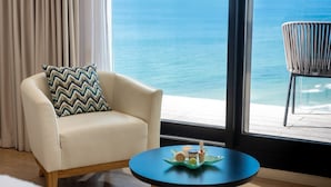 A room with a view of the ocean, a white armchair with a patterned pillow, a small blue table with a decorative item, and a wicker chair by the window. - Vert Lagoon Netanya By AFI Hotels (Netanya)