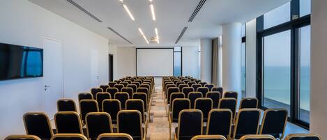 A conference room with rows of chairs facing a projection screen, large windows on one side, and a flat-screen TV on the wall.