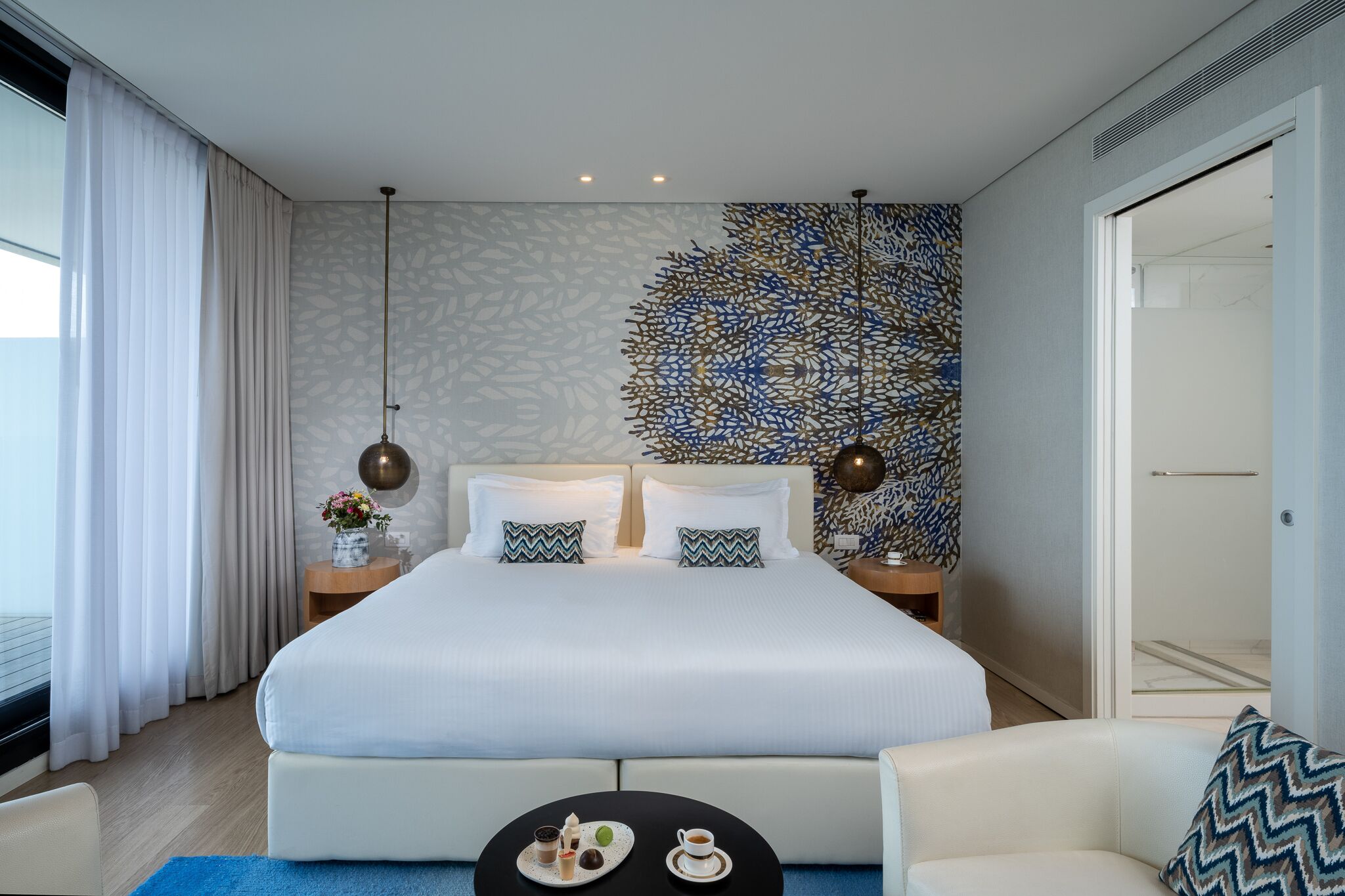 A modern hotel room with a large bed, two nightstands, a coffee table, and a view of the sea.