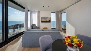 A modern living room with a grey sofa, a small table with a vase of flowers, and a view of the sea through large windows. - Vert Lagoon Netanya By AFI Hotels (Netanya)