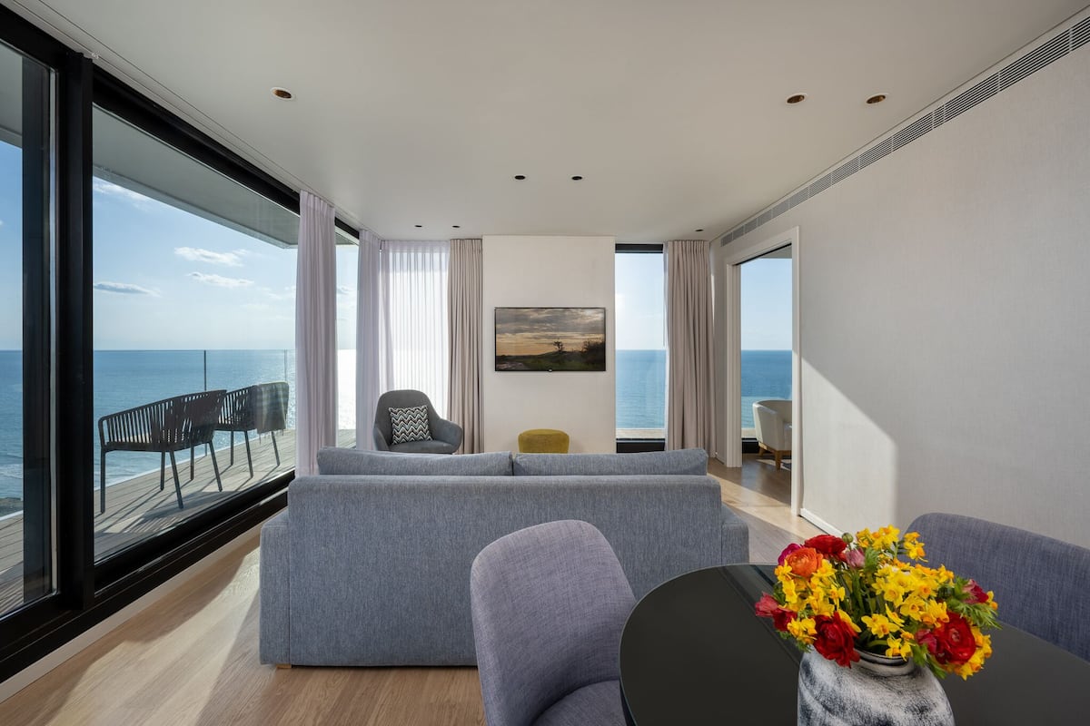 A modern living room with a grey sofa, a small table with a vase of flowers, and a view of the sea through large windows.