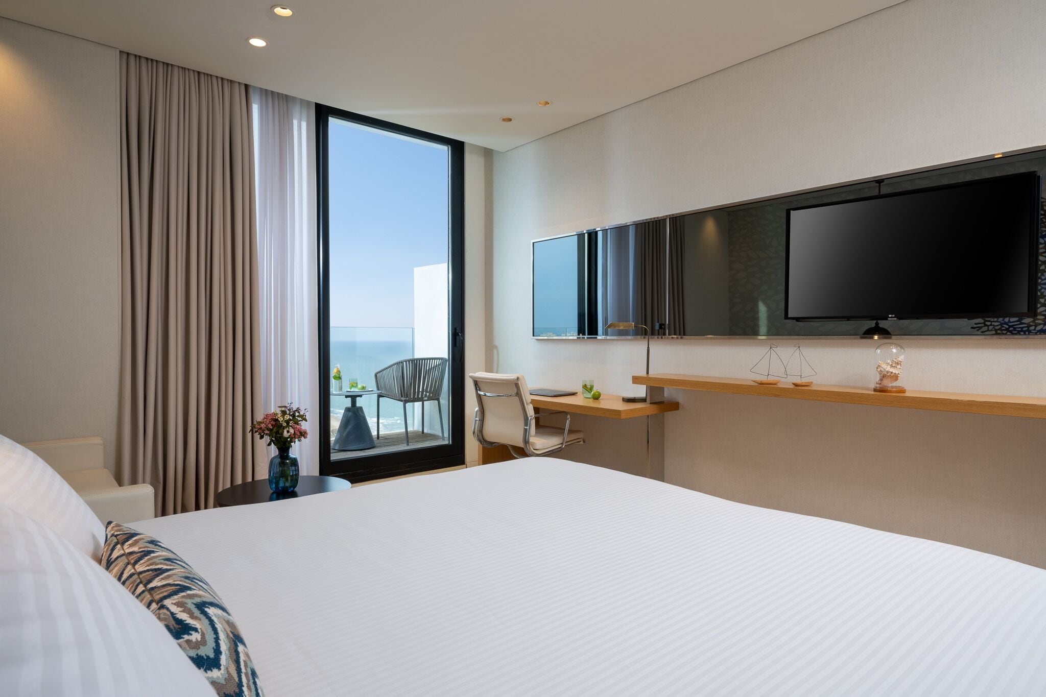 A hotel room with a large bed, a desk with a chair, a flat-screen TV mounted on the wall, and a window with curtains.