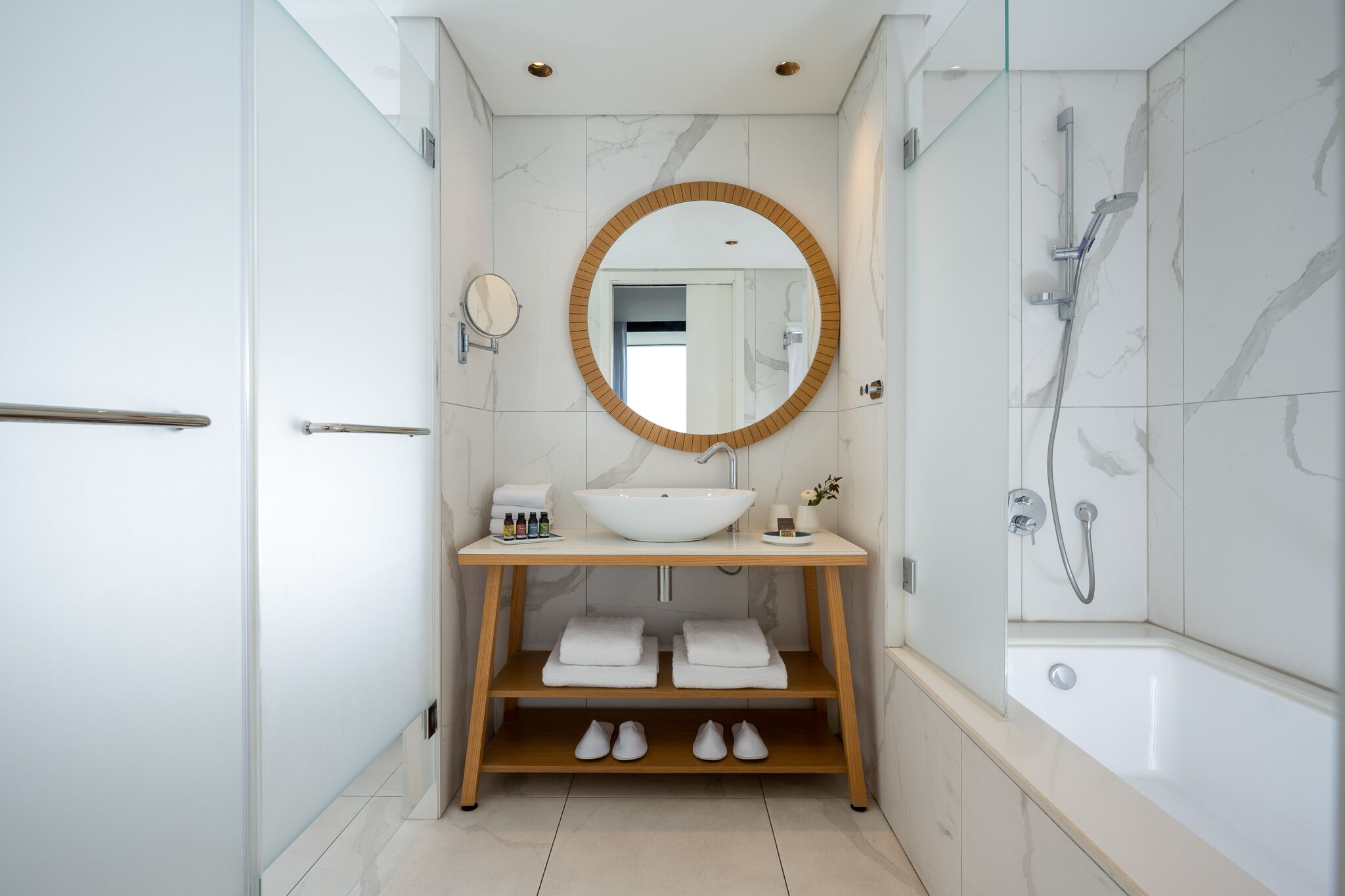 A modern bathroom with a white sink, wooden vanity, a round mirror, and a shower area.