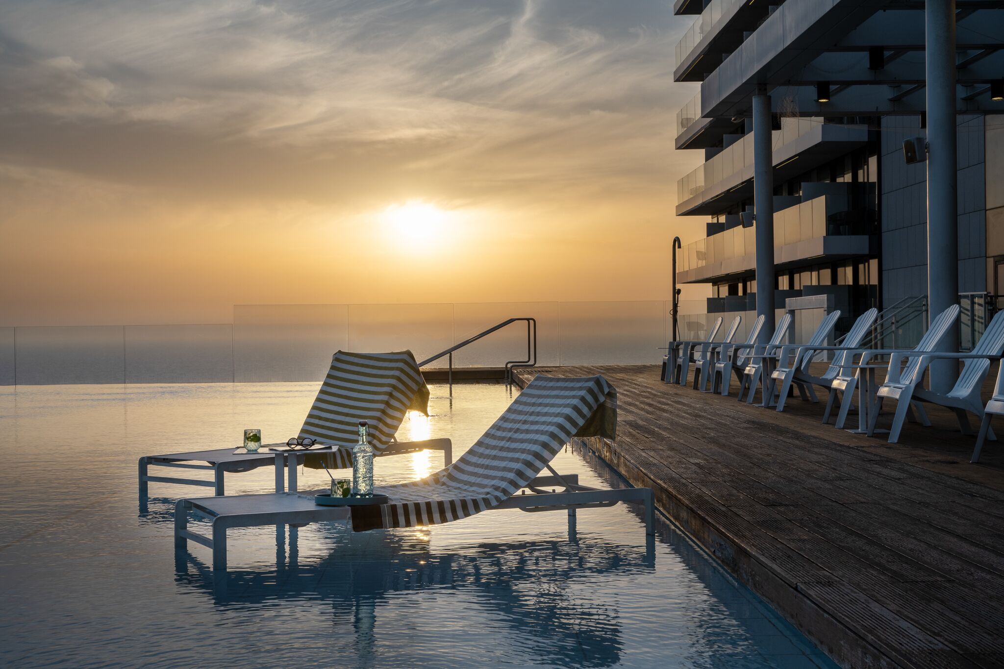 A poolside area with lounge chairs and tables, overlooking a body of water with a sunset in the background.