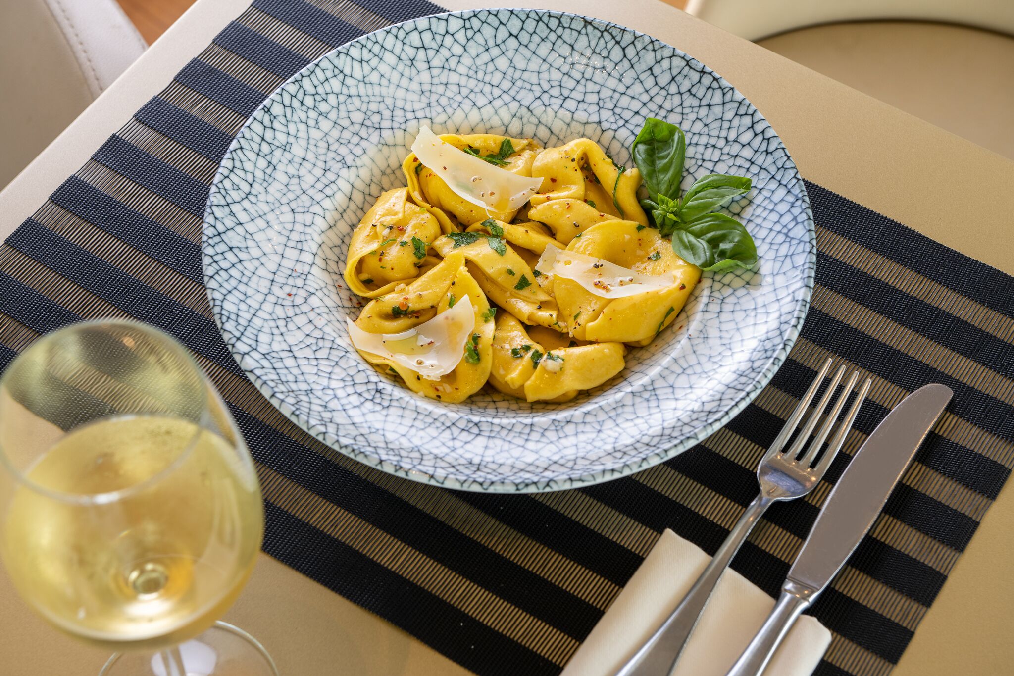 Un plato de pasta con un adorno de albahaca, una copa de vino blanco y cubiertos sobre un mantel a rayas.