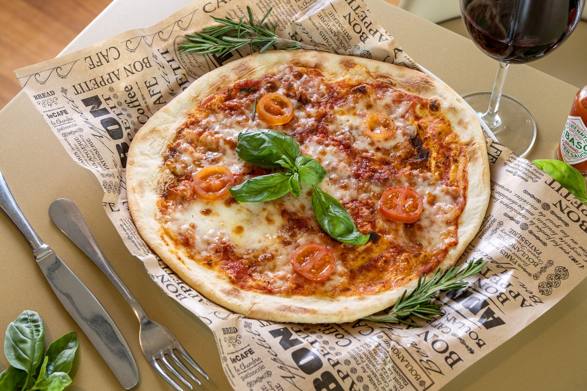 Una pizza sobre una mesa forrada con periódico, junto a una copa de vino tinto, un tenedor y un cuchillo.