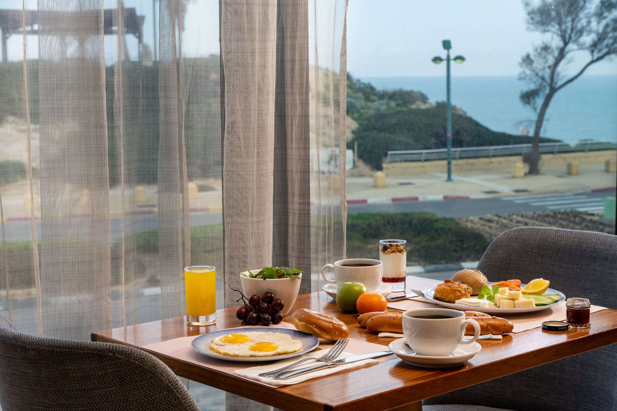 A breakfast tray with eggs, toast, fruit, and coffee on a table by a window overlooking a coastal view.