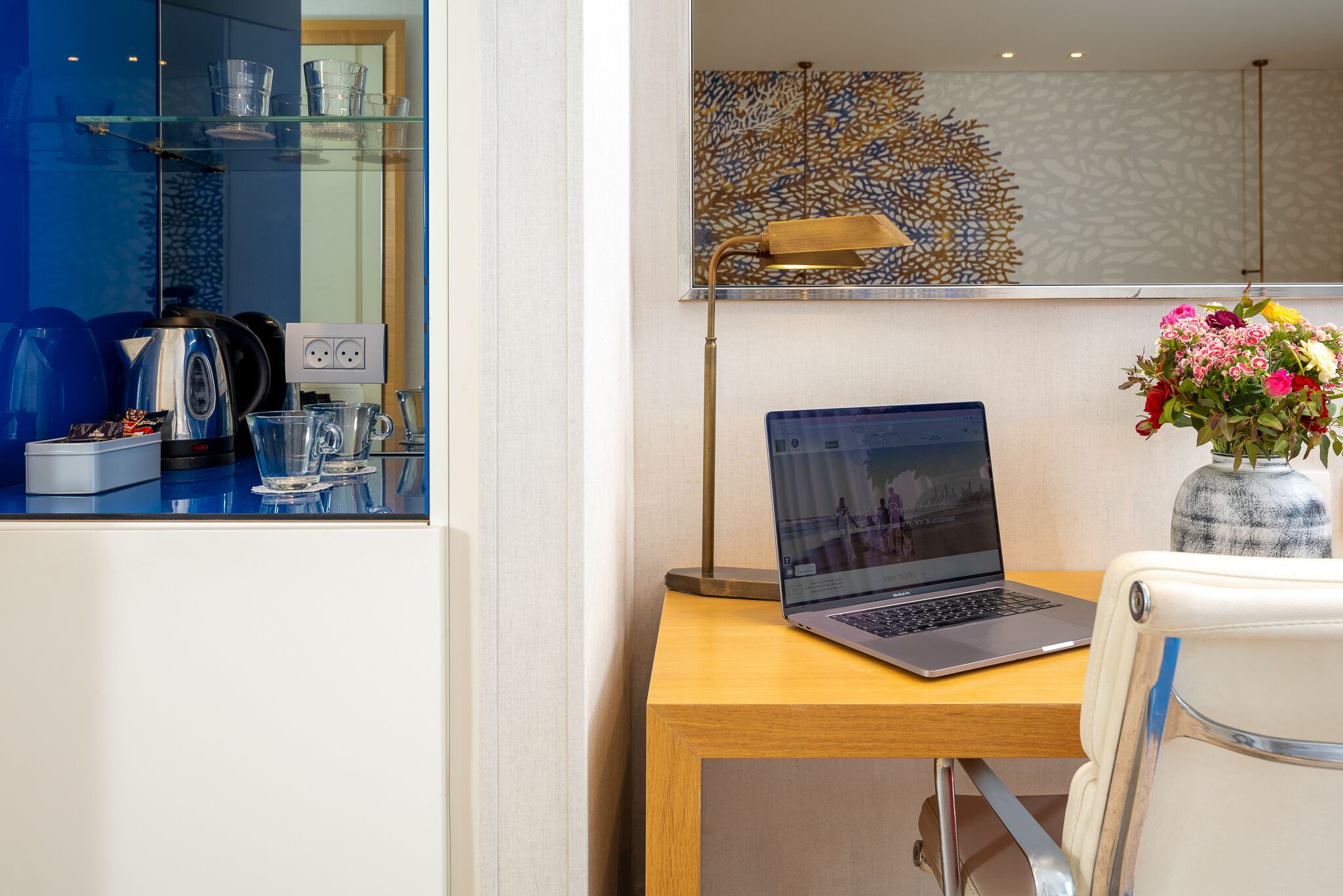 Ein modernes Zimmer mit einem Laptop auf einem Holztisch, einer Vase mit Blumen und einer blauen Glasschrankwand mit Küchenutensilien.