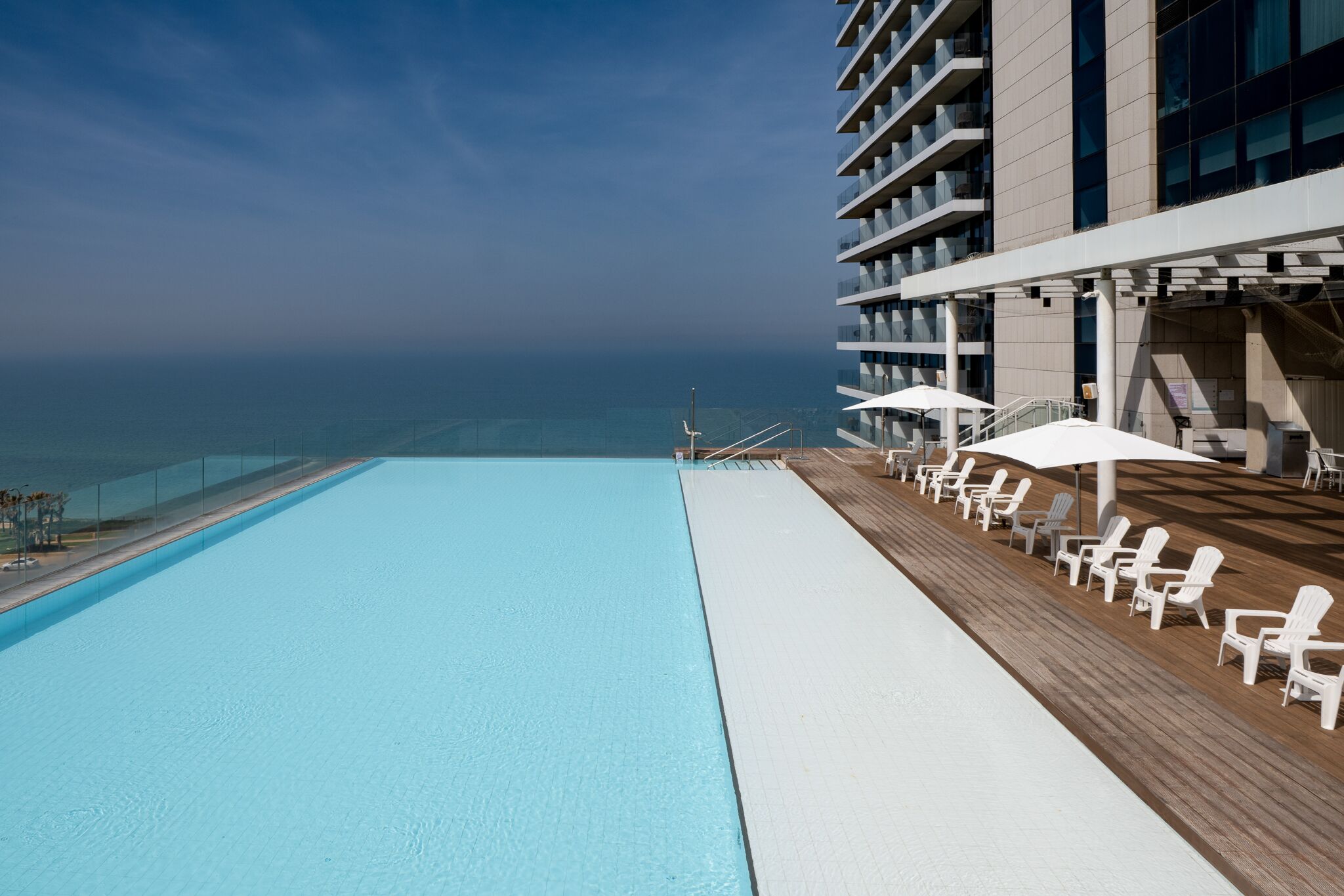 A rooftop pool with a view of the ocean, surrounded by white lounge chairs and umbrellas.