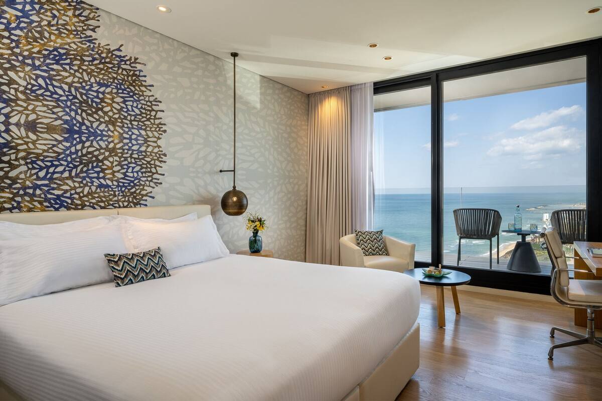 A modern hotel room with a large bed, a view of the sea, and a dining area with chairs and a table.