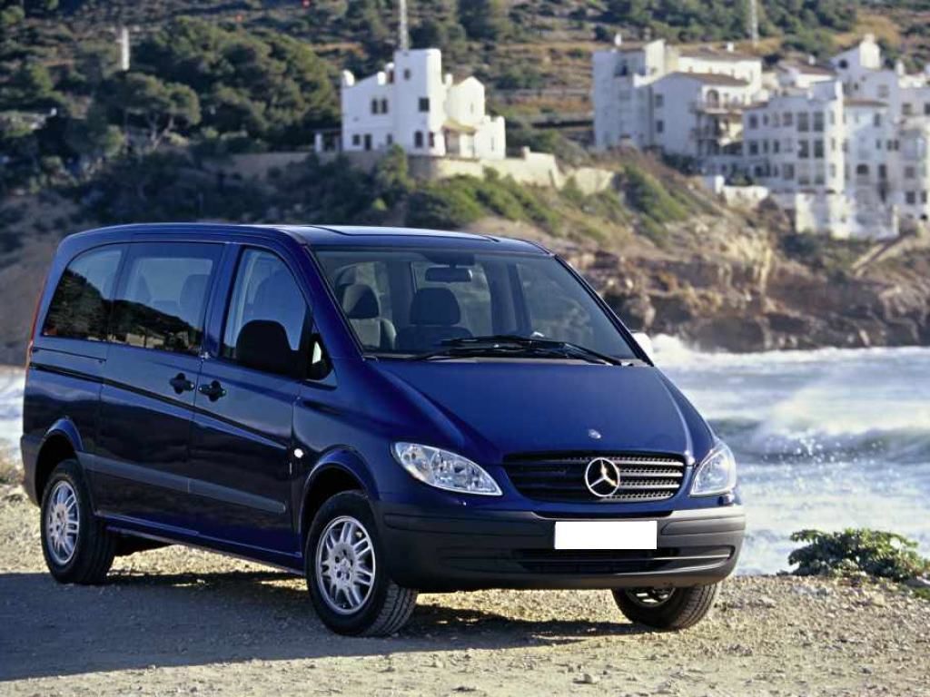 A blue car parked on a beach with white buildings in the background.