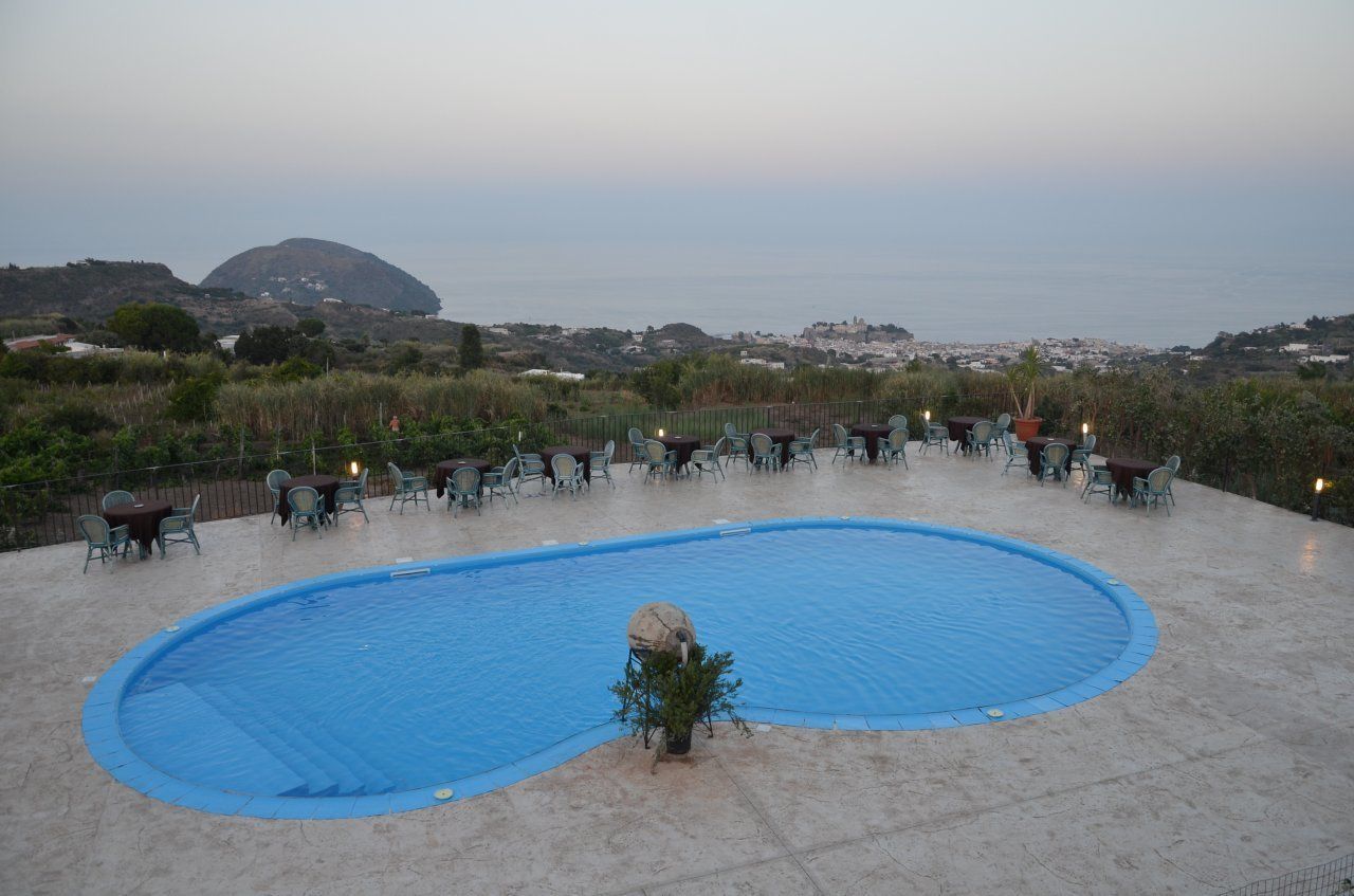 A pool area with a blue pool, surrounded by outdoor seating and a view of the sea and hills.