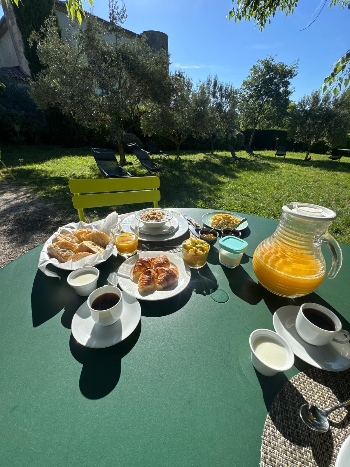 An outdoor dining table with breakfast items, including croissants, fruit, and coffee, set against a backdrop of a green lawn and a building.