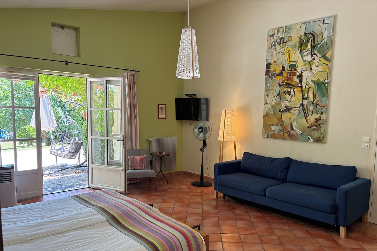 Une pièce comprenant un lit, un canapé bleu, un ventilateur sur pied, une table et un tableau accroché au mur.