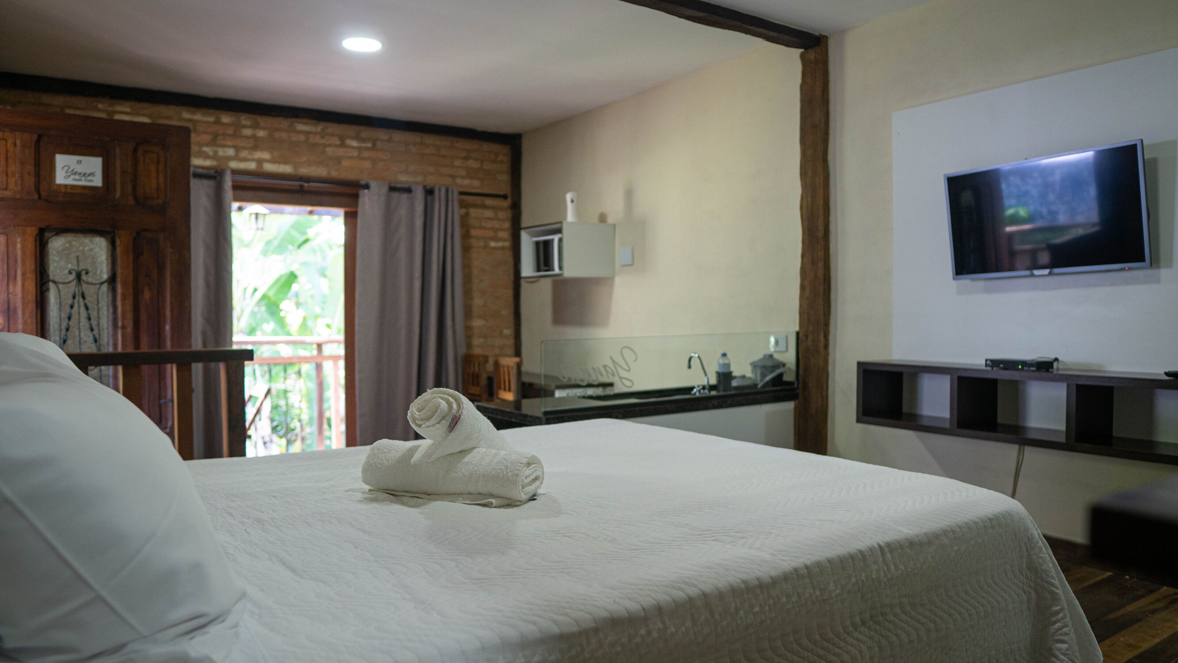 A hotel room with a bed, a TV mounted on the wall, a small kitchen area, and a balcony with curtains.