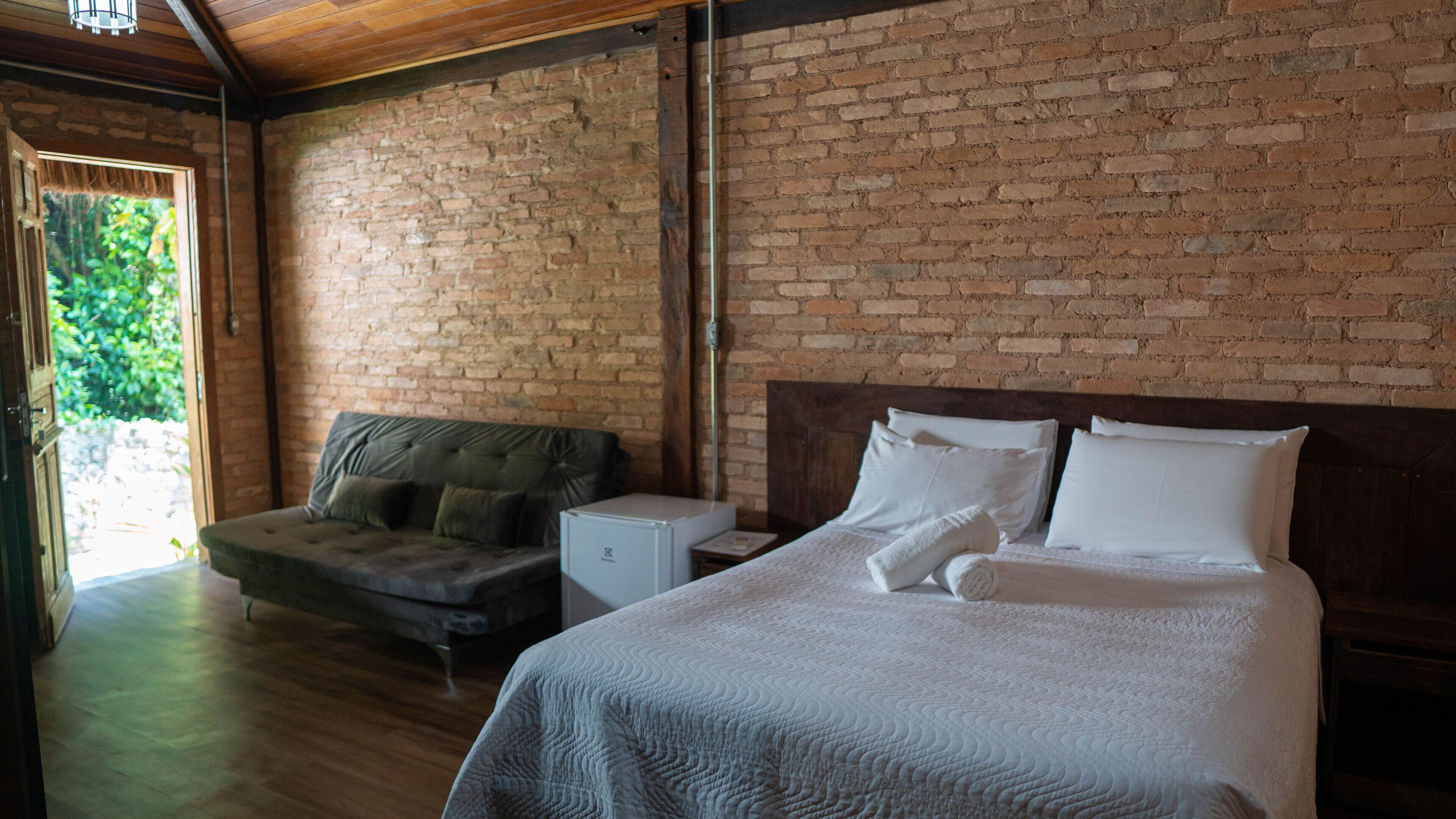 Un dormitorio con pared de ladrillo, una cama con ropa de cama blanca, cabecera de madera y un sofá con cojines.