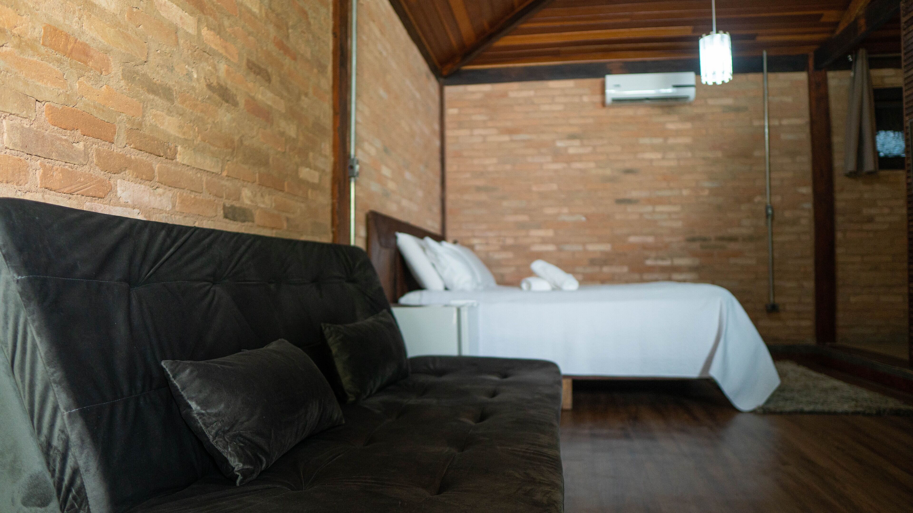 Una habitación con un sofá marrón oscuro, una cama blanca con almohadas y una pared de ladrillo.