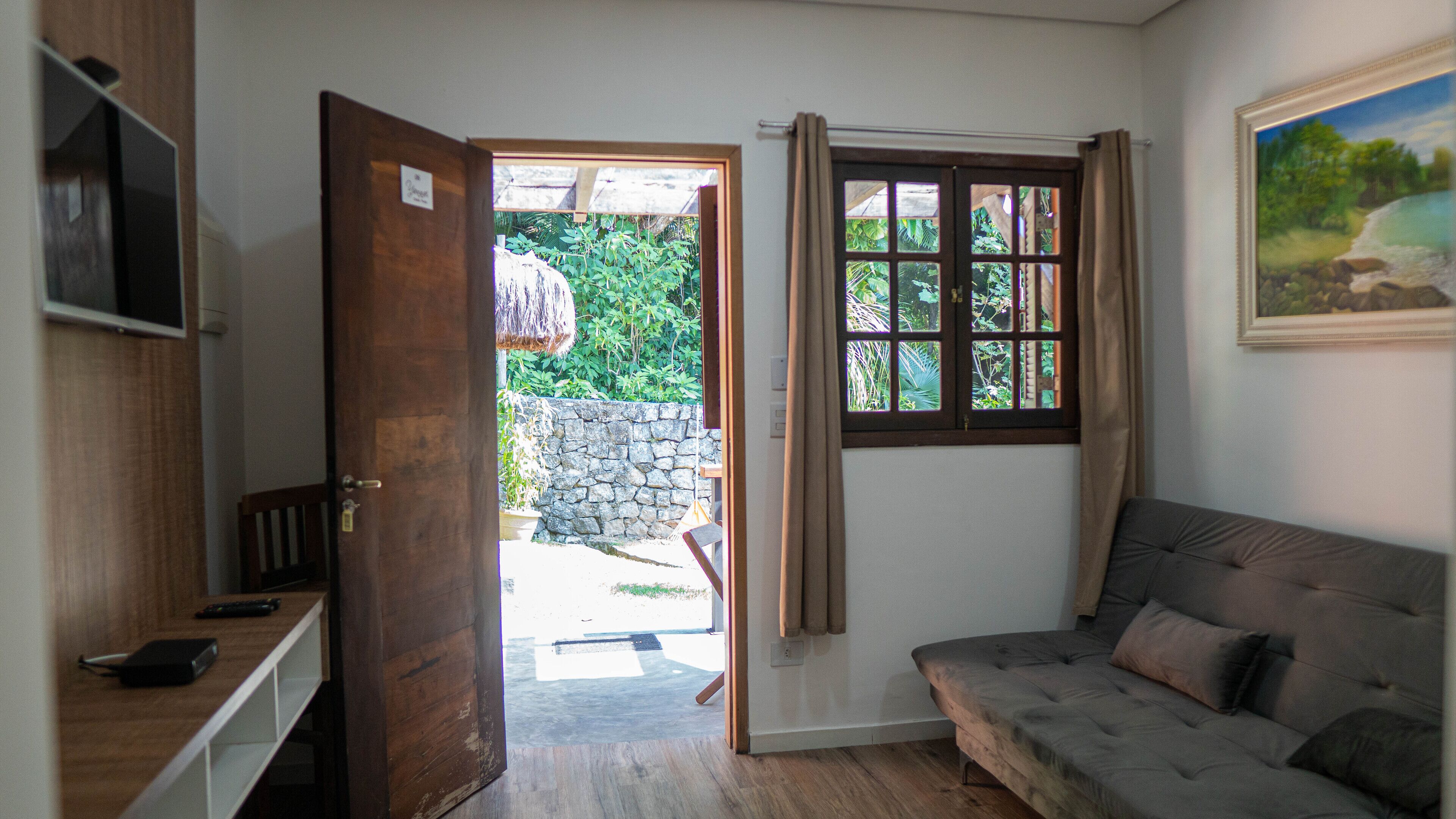 Une pièce avec une porte en bois donnant sur un espace extérieur, une fenêtre avec des rideaux, un canapé gris et un tableau accroché au mur.
