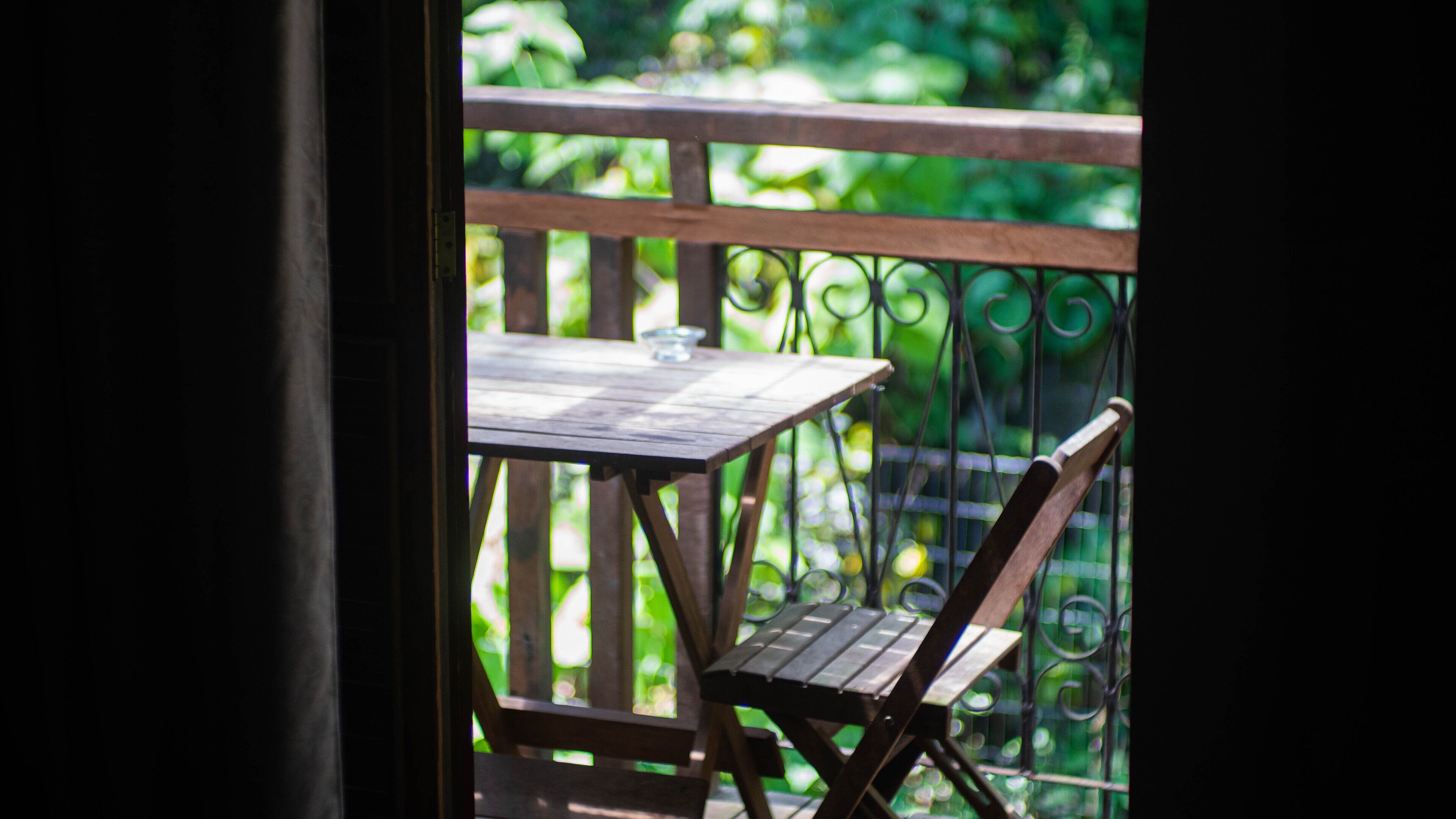 A wooden table and chair on a balcony with a metal railing, overlooking a garden.