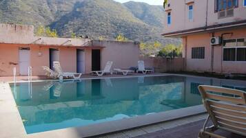 Piscina com espreguiçadeiras, um edifício com várias entradas e montanhas ao fundo.